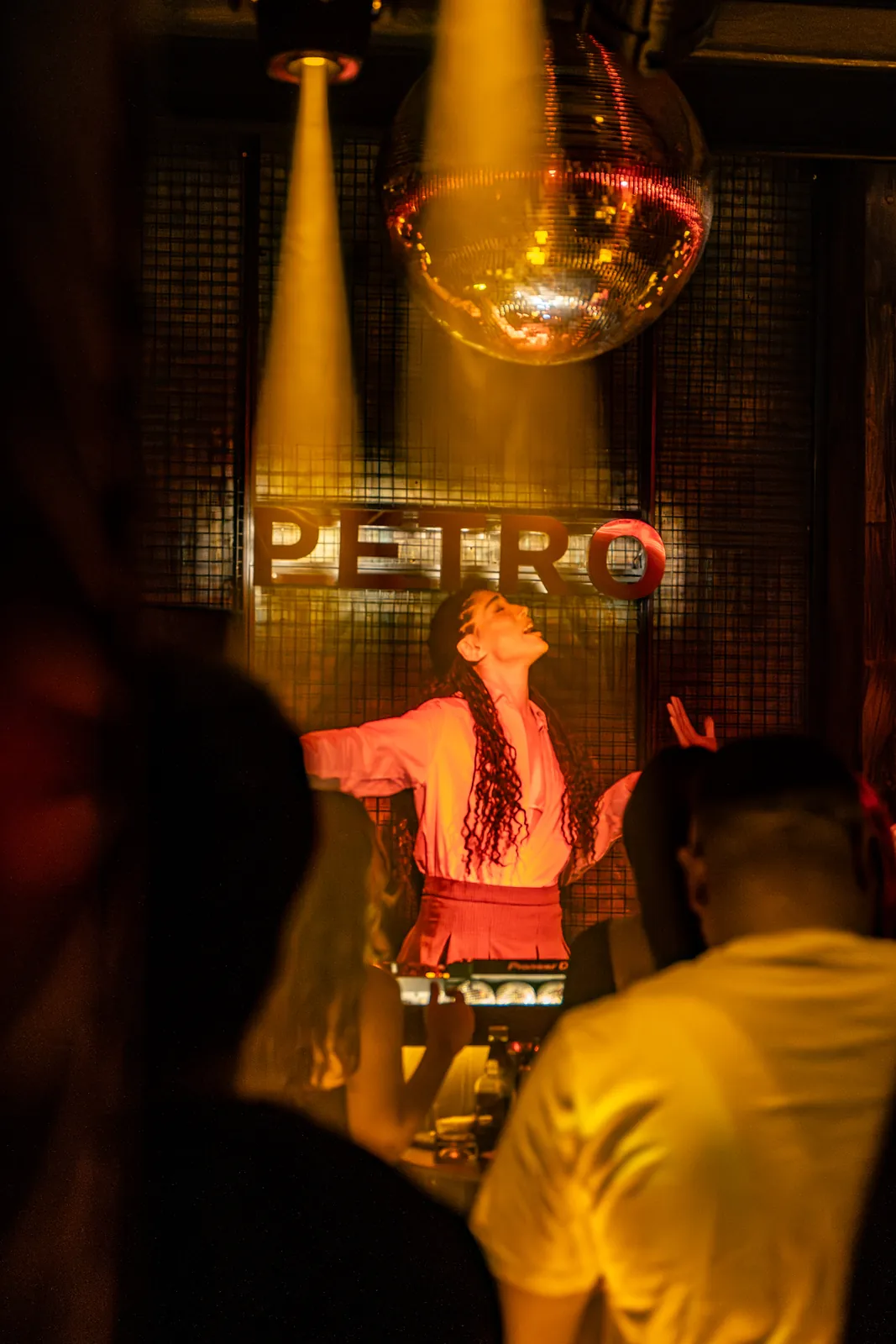 Petro Cocktail Bar — A woman DJ under a yellow spotlight and the disco ball, the crowd in the foreground