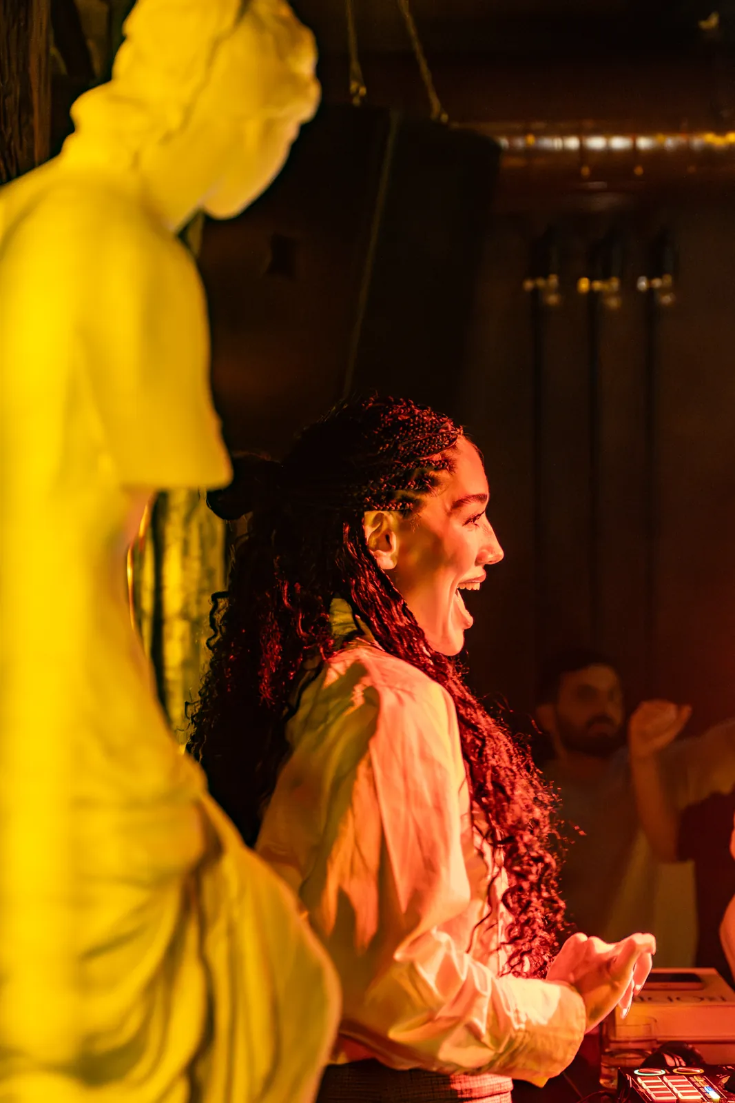 Petro Cocktail Bar — A classical statue in yellow light beside a smiling woman DJ
