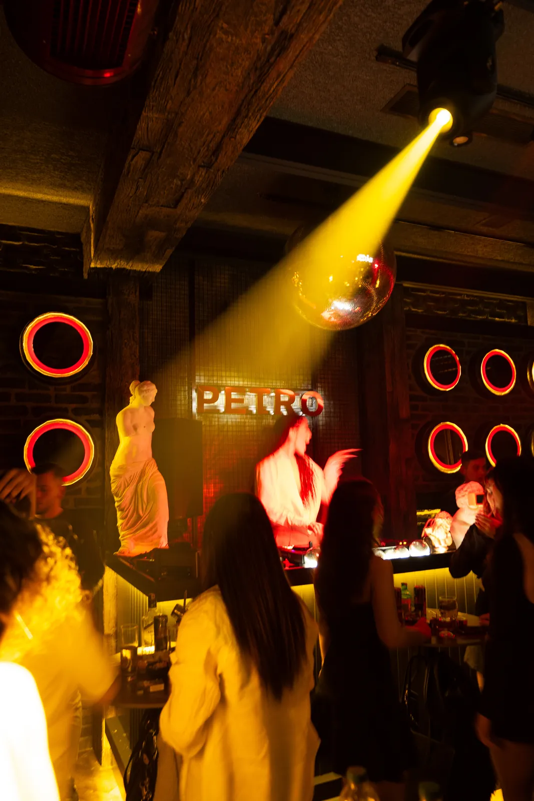 Petro Cocktail Bar — A yellow light beam, statues and the DJ at the booth, guests dancing in the foreground